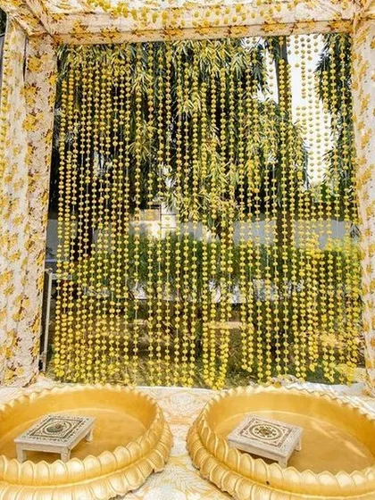 This intimate haldi seating is framed by curtains made of yellow floral prints and strings of marigolds. The setup is perfect for home ceremonies, creating a beautiful and sacred space with minimal elements.