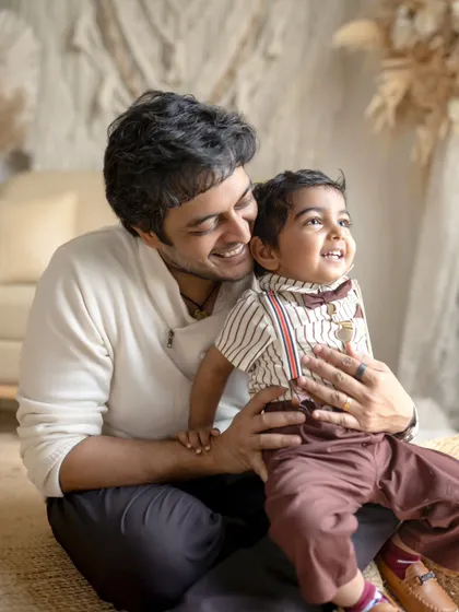 A father and son share a happy, candid moment. The boy is sitting on his father's lap, and they are both looking off-camera with bright smiles.