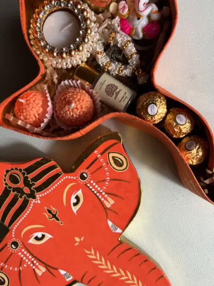 A close-up of the items inside the Ganesha box, including motichoor laddoos, Ferrero Rocher, a mini perfume, and a pearl-studded candle holder.