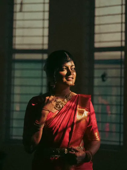 A moody and atmospheric portrait of a bride. The shadows from the window create a dramatic effect, highlighting the texture of her red saree and her serene expression.
