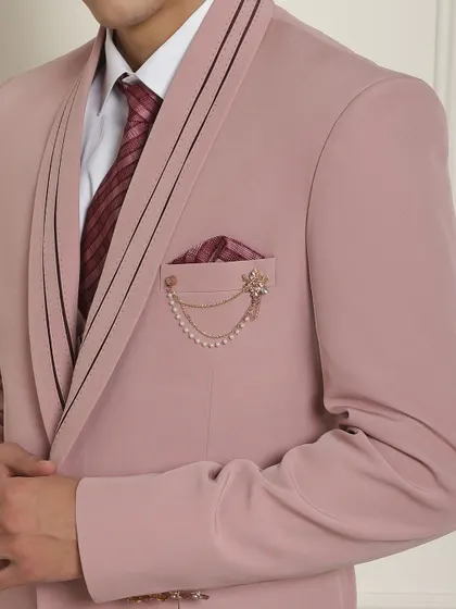 A detailed view of a pink suit's lapel, showcasing the fine stitching and a pearl chain accessory. It’s these touches that define our tailoring.