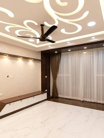 An ornate false ceiling with a swirling design adds a touch of drama to this living room. The dark wood entertainment unit and shelving provide a beautiful contrast against the white marble-finish floor.