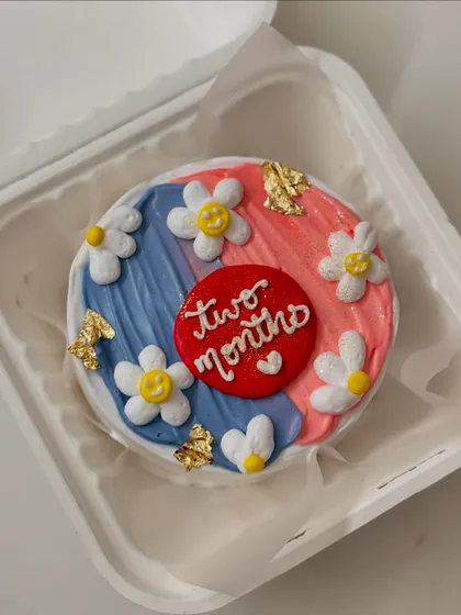 A sweet and cheerful bento cake for a two-month celebration. The cake has a pink and blue background, decorated with smiling daisies and touches of gold leaf.