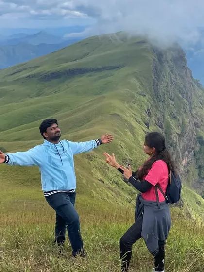 A fun, candid moment between two trekkers with the stunning Nethravathi peak in the background.