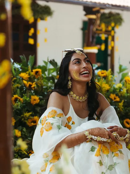 The bride, with a smear of turmeric on her cheek, looks up with a radiant smile. Her floral-printed outfit and the sunflower-filled background create a picture of pure happiness.