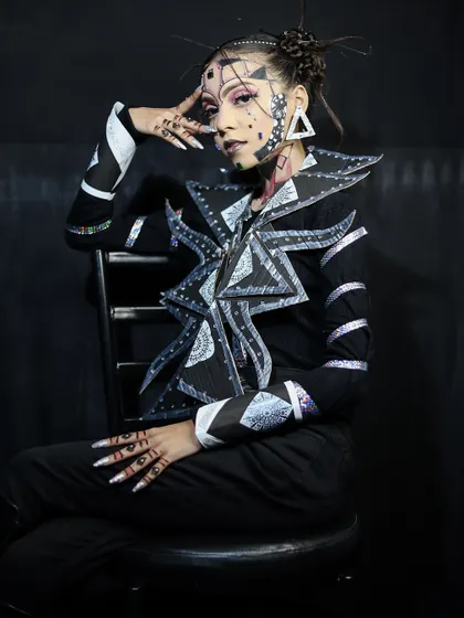 A full-length seated pose, showing the complete avant-garde outfit and makeup.