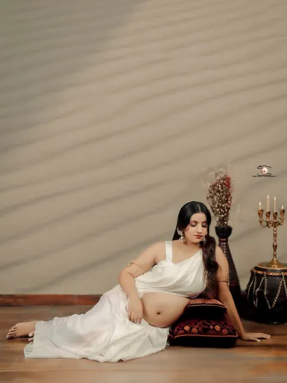 A peaceful, reclining pose of the mother-to-be in a white saree. Lying on a floor cushion, she cradles her bump, with the soft studio lighting creating a calm and intimate atmosphere.