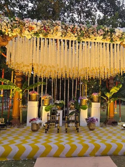 A stunning South Indian theme Haldi stage. The mandap is built with traditional wooden pillars, a canopy of jasmine flowers, and a vibrant yellow and white chevron-patterned floor.