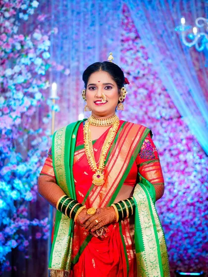A classic portrait of a Maharashtrian bride in a red and green saree. She is adorned with traditional gold jewellery, including a long necklace and nath, against a festive floral background.