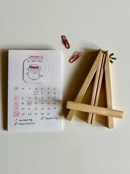 The full 2025 desk calendar set, showing the January page, the wooden easel, and the red clips used to hold the pages in place.