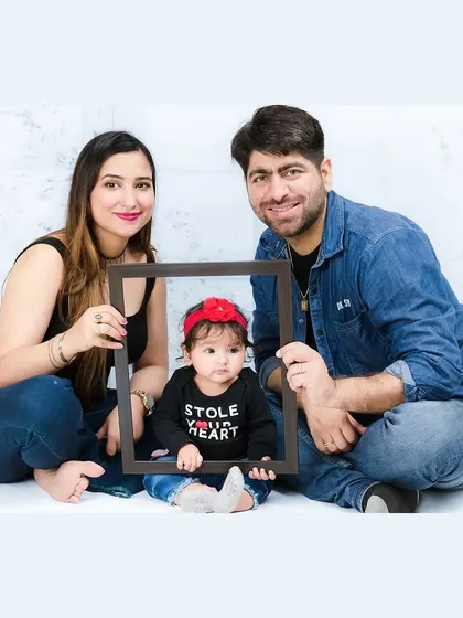 A creative family portrait where the parents hold a frame around their adorable baby, who is wearing a "Stole Your Heart" t-shirt. It's a fun and modern take on the family photo.