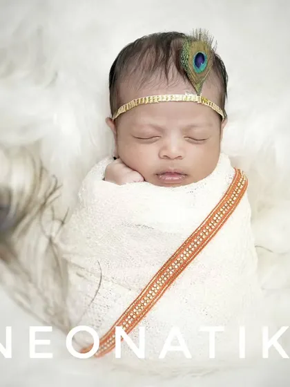 A minimalist and modern take on the newborn Krishna theme, with the baby wrapped in white on a fluffy surface, accented by a single peacock feather.