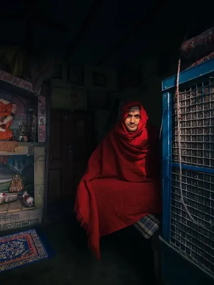 A person wrapped in a red blanket sits inside a modest room, the low light creating a moody and intimate indoor portrait.