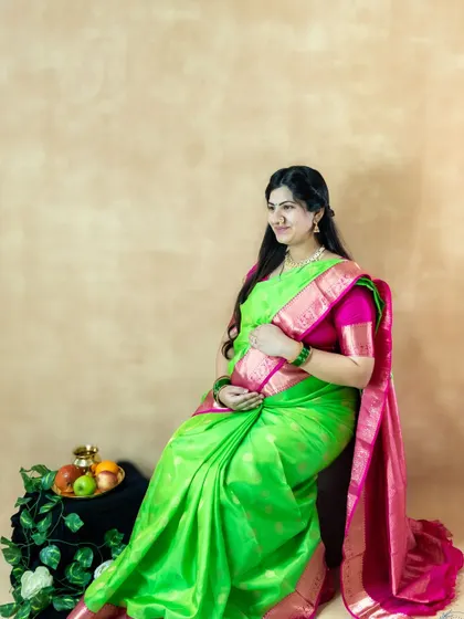 A beautiful studio portrait of a mom-to-be in a green and pink saree, seated with a traditional fruit offering.