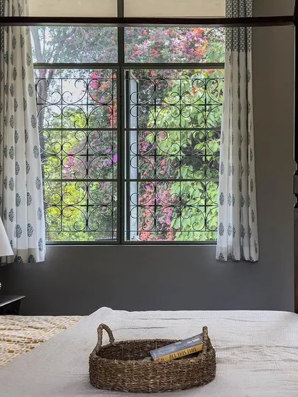 This bedroom is designed as a serene retreat, with a classic four-poster bed and large windows. The windows feature traditional-style grilles and are framed by block-printed curtains, overlooking the garden.