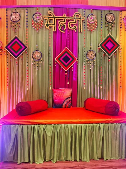 For a Mehendi night, I can create a stunning backdrop like this one. It includes a comfortable diwan for the bride, decorated with colourful drapes, traditional hangings, and Rajasthani umbrellas for beautiful photos.