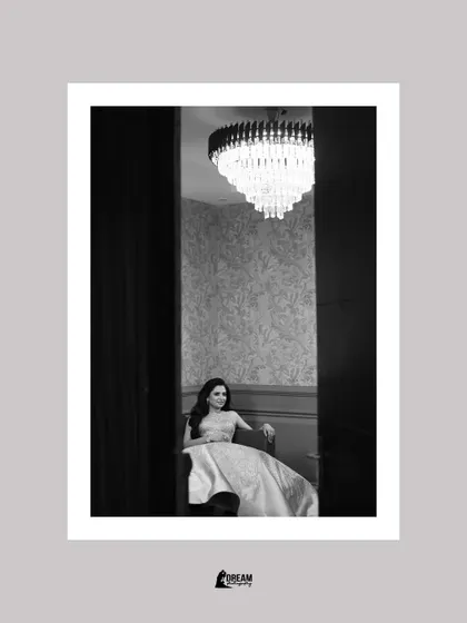 A peek-a-boo shot of bride Harshna, seated elegantly under a chandelier at her Sagan ceremony.