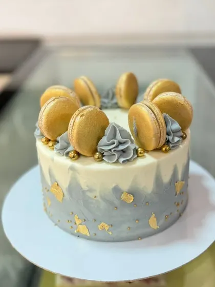 A stylish grey and white watercolor cake. This design is elegantly decorated with gold leaf, grey buttercream swirls, and a crown of golden macarons.