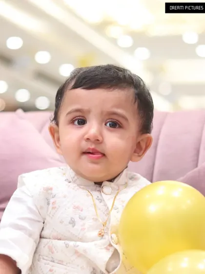 A close-up portrait of a baby boy in a formal vest, holding a yellow balloon.