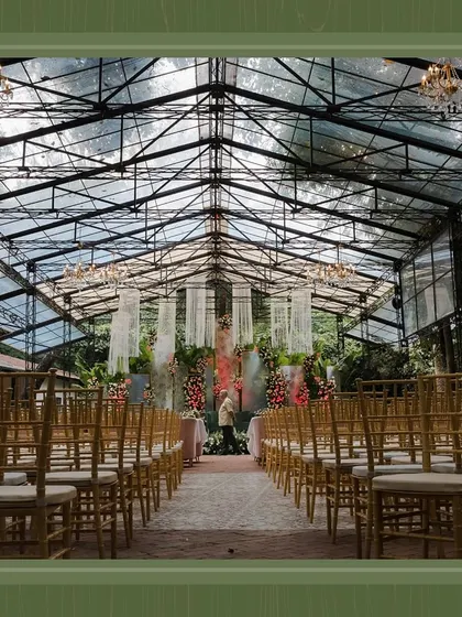 A view down the aisle towards the stage, showing the guest seating arrangement. The glass ceiling allows natural light to flood the space, highlighting the floral decor.