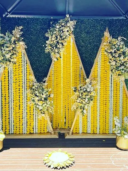 A stunning Haldi backdrop for a deck or patio. The triangular panels are covered in marigold strings and accented with white flowers, creating a modern and elegant design.
