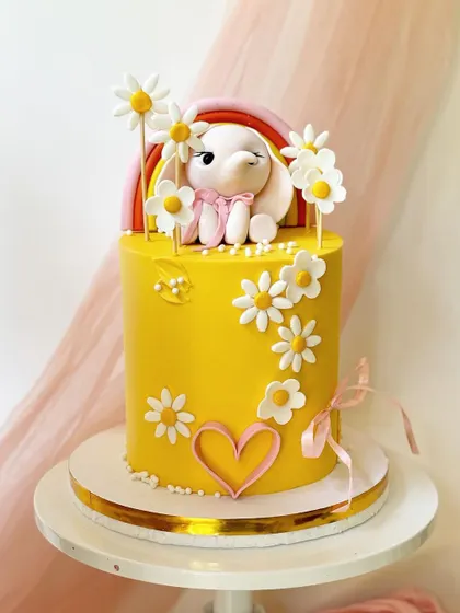 A sunny yellow cake with a daisy theme. The design includes a cute bunny topper, a rainbow accent, and a piped heart outline.