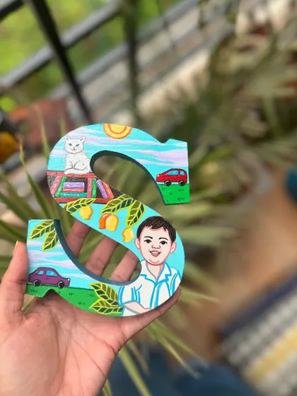 A custom wooden letter 'S' painted for a child, featuring his portrait, his cat, and his favorite toys.