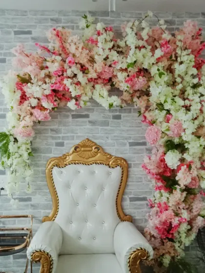 A simple yet elegant photo corner featuring a white throne chair set against a brick wall. The asymmetrical arch of artificial pink and white cherry blossoms adds a soft, beautiful touch.