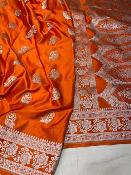 A bright orange Mashru silk saree that is perfect for festive celebrations. The silver floral weaving adds a touch of traditional elegance to the vibrant color.