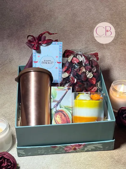 Shine bright with our Diwali delights. This elegant hamper includes a copper tumbler, a layered candle, a bag of potpourri, and a box of delicious chocolates, all in a beautiful floral box.