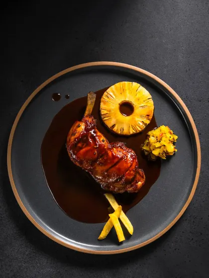 A gourmet dish of grilled chicken leg with a pineapple ring in a dark sauce, shot from above for a dramatic effect.