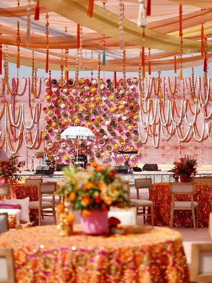 Welcome to a 'Phulkari Fasana' or a floral story. This vibrant Mehendi setup features a stunning floral wall, traditional embroidered fabrics, and a lively color palette of pinks and oranges.