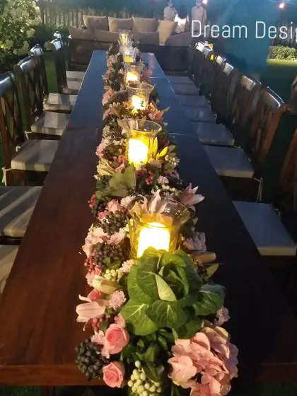 A long wooden table set for an outdoor dinner. The centerpiece is a continuous runner of lush flowers, greenery, and candles in hurricane lamps, creating a rustic yet romantic look.
