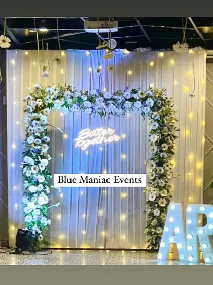 A modern and chic engagement backdrop with a square floral frame of white flowers. The 'Better Together' neon sign, marquee letters, and hanging lights create a fun and stylish photo opportunity.