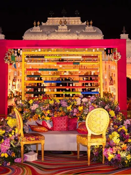 A vibrant and colorful seating area for a Mehendi, with a unique backdrop made of colored spools and an abundance of fresh flowers, set against the grandeur of Shiv Niwas Palace.
