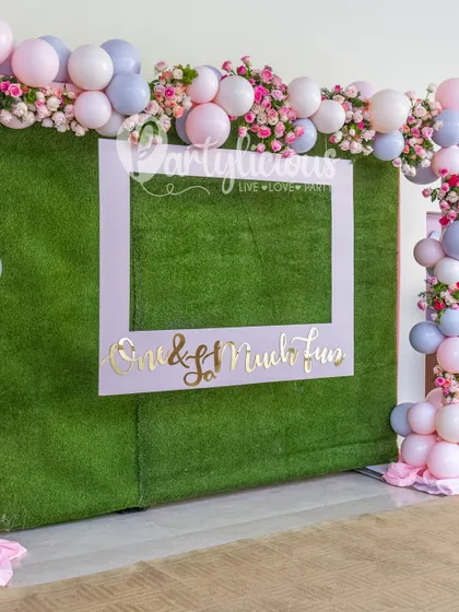 A fun and interactive photo frame against a green hedge wall, decorated with pink and lilac balloons.