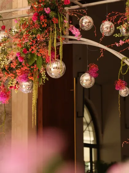 A close-up of the "Retro Disco" Mehendi entrance arch, showing hanging disco balls nestled within vibrant pink and orange floral arrangements.