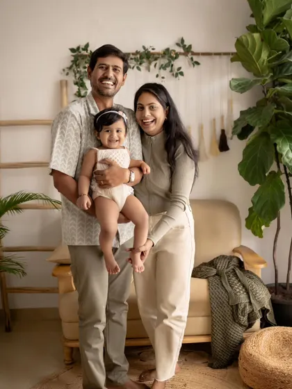 A joyful family of three, with the parents holding their smiling daughter. The natural textures and indoor plants create a warm and stylish backdrop.