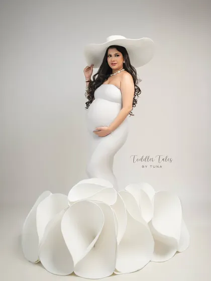 A vision in white. This couture gown, with its sculptural flower petal skirt, transforms the mother-to-be into a work of art. The wide-brimmed hat adds a final touch of classic Hollywood glamour.