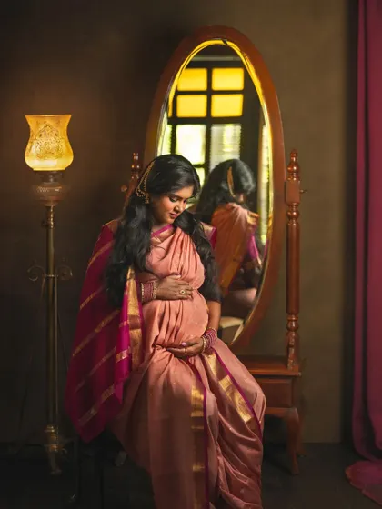 A duplicate of ID 46, this beautiful studio portrait shows an expecting mother in a traditional silk saree, seated before a vintage mirror. The warm light from the lamp adds a golden glow, creating a rich, classic composition.