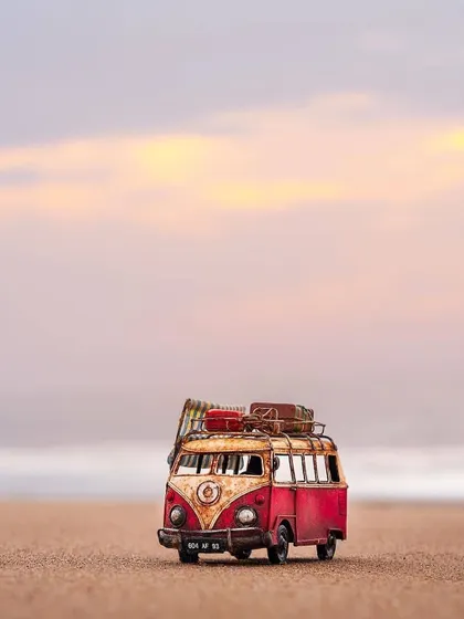 A miniature, rustic model of a Volkswagen bus sits on a sandy beach against a soft sunset sky. This image evokes feelings of wanderlust and vintage travel.