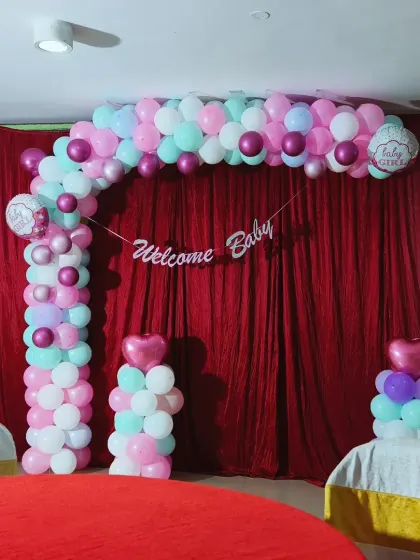 A simple and sweet "Welcome Baby" decoration for a baby girl's naming ceremony or homecoming party. The pastel balloon arch against a red curtain provides a lovely pop of color.