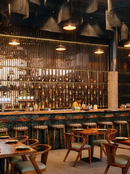 A frontal view of the main bar, the heart of the restobar. The design features a live edge wood countertop, green subway tiles, and a dramatic, back-lit bottle display. The expanded metal mesh ceiling above the bar adds another layer of industrial texture.