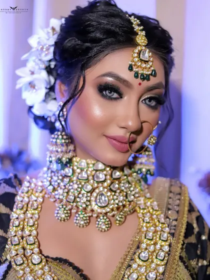 A stunning close-up of a bride in a black and gold lehenga, adorned with white flowers in her hair. The focus is on her dramatic smokey eye makeup and heavy kundan jewellery.