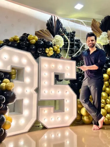 This 35th birthday decor uses a similar concept with large light-up numbers, but with added details like black and gold palm leaves for a more stylized, art-deco feel. The client is pictured enjoying the setup.