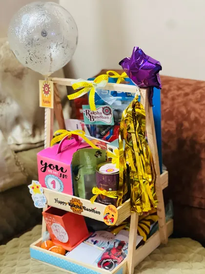 A tall, two-tiered wooden stand hamper for Rakhi, decorated with balloons and filled with a variety of colorful gifts.