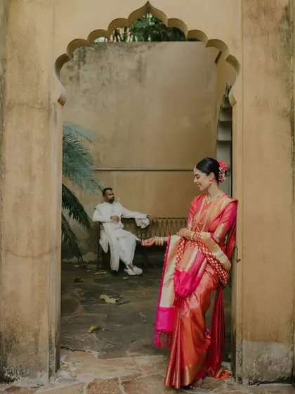 An artistic shot from a Sri Lankan wedding. The bride, in her vibrant Kanjeevaram sari, stands in an archway while the groom waits in the background, creating a sense of anticipation and beautiful composition.