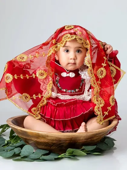 A baby girl in a beautiful red traditional outfit with a veil sits in a wooden bowl, a unique and culturally rich portrait.