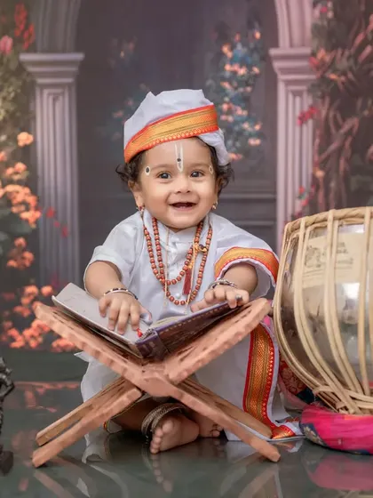 A happy baby Varkari, seated with a holy book and tabla against a beautiful floral backdrop. This image blends tradition with a touch of modern studio artistry.