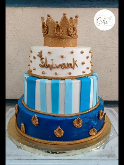 A royal three-tier cake fit for a little prince. This design features a bold blue and white color scheme with stripes, gold accents, and a glittering gold crown topper.